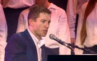 Lee Evans sings at the piano with a choir backing